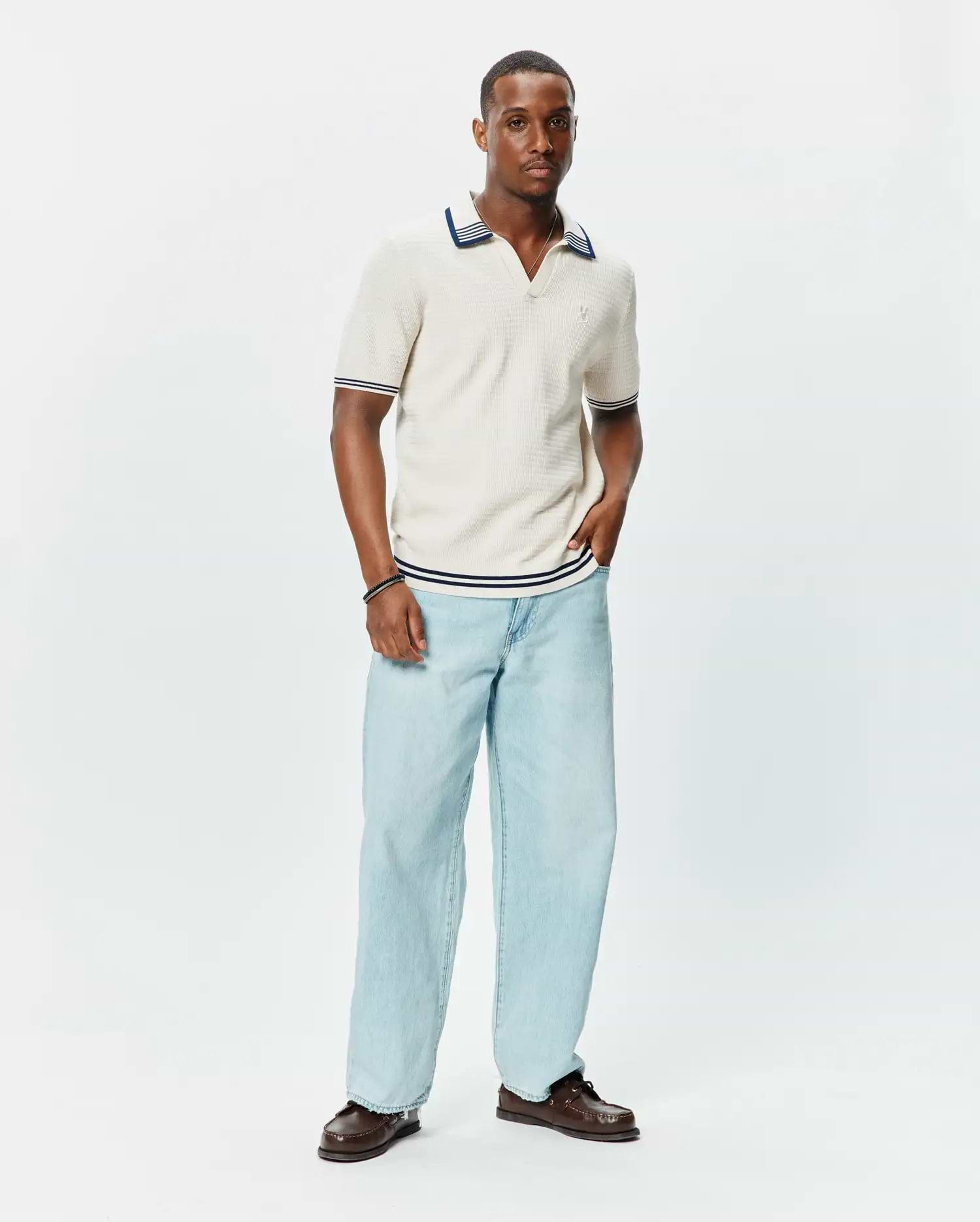 Mens Elowen Knit Polo in cream, featuring a ribbed texture, V-neck, and navy blue stripe accents on the collar, cuffs, and hem, with a small embroidered Psycho Bunny logo on the chest. (3)