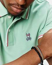 Mens Faye Pique Polo in mint green, featuring a classic fit and pique knit fabric. It has a small embroidered Psycho Bunny skull logo on the left chest, with white and pink striped collar and sleeve cuffs.
