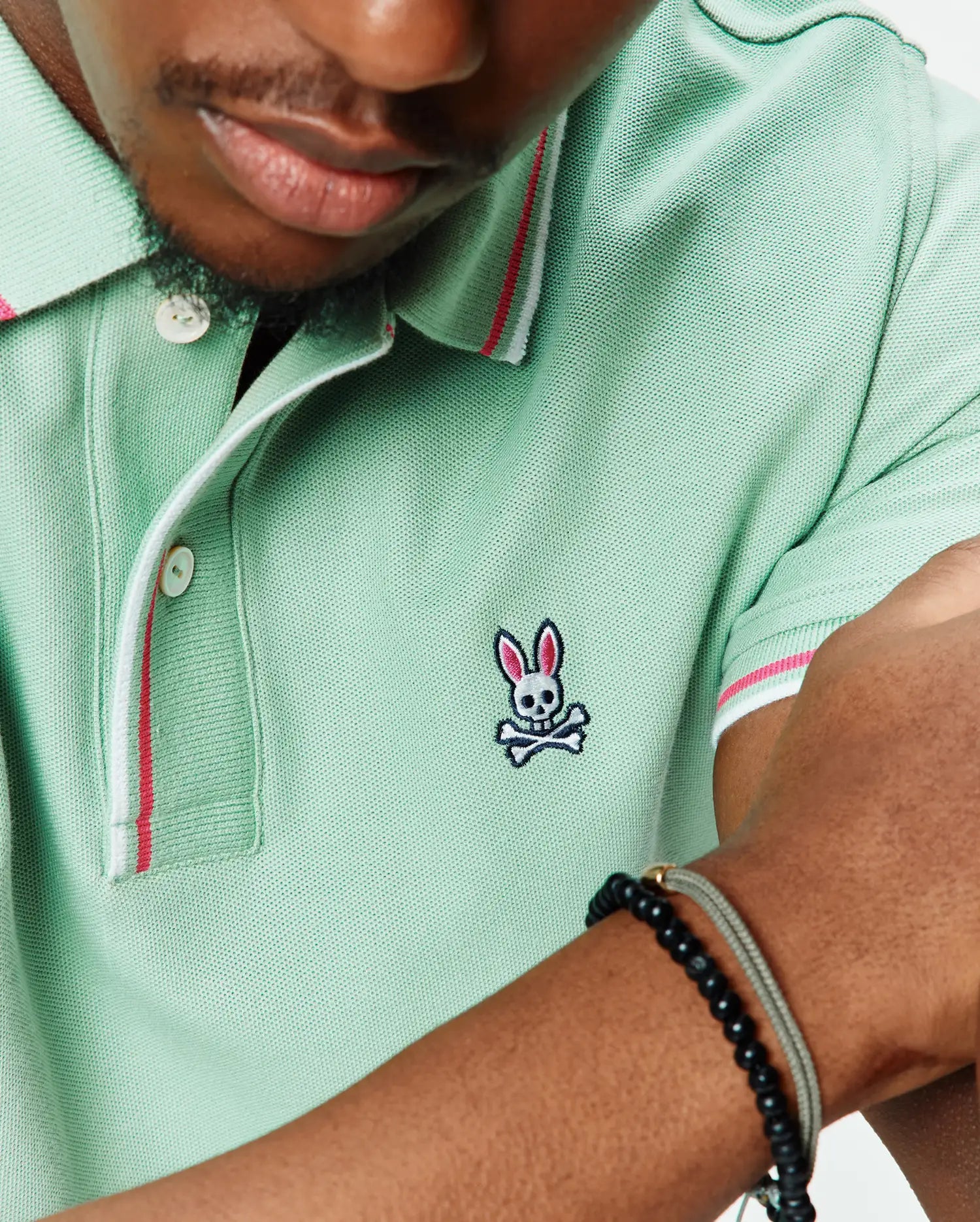 Mens Faye Pique Polo in mint green, featuring a classic fit and pique knit fabric. It has a small embroidered Psycho Bunny skull logo on the left chest, with white and pink striped collar and sleeve cuffs.