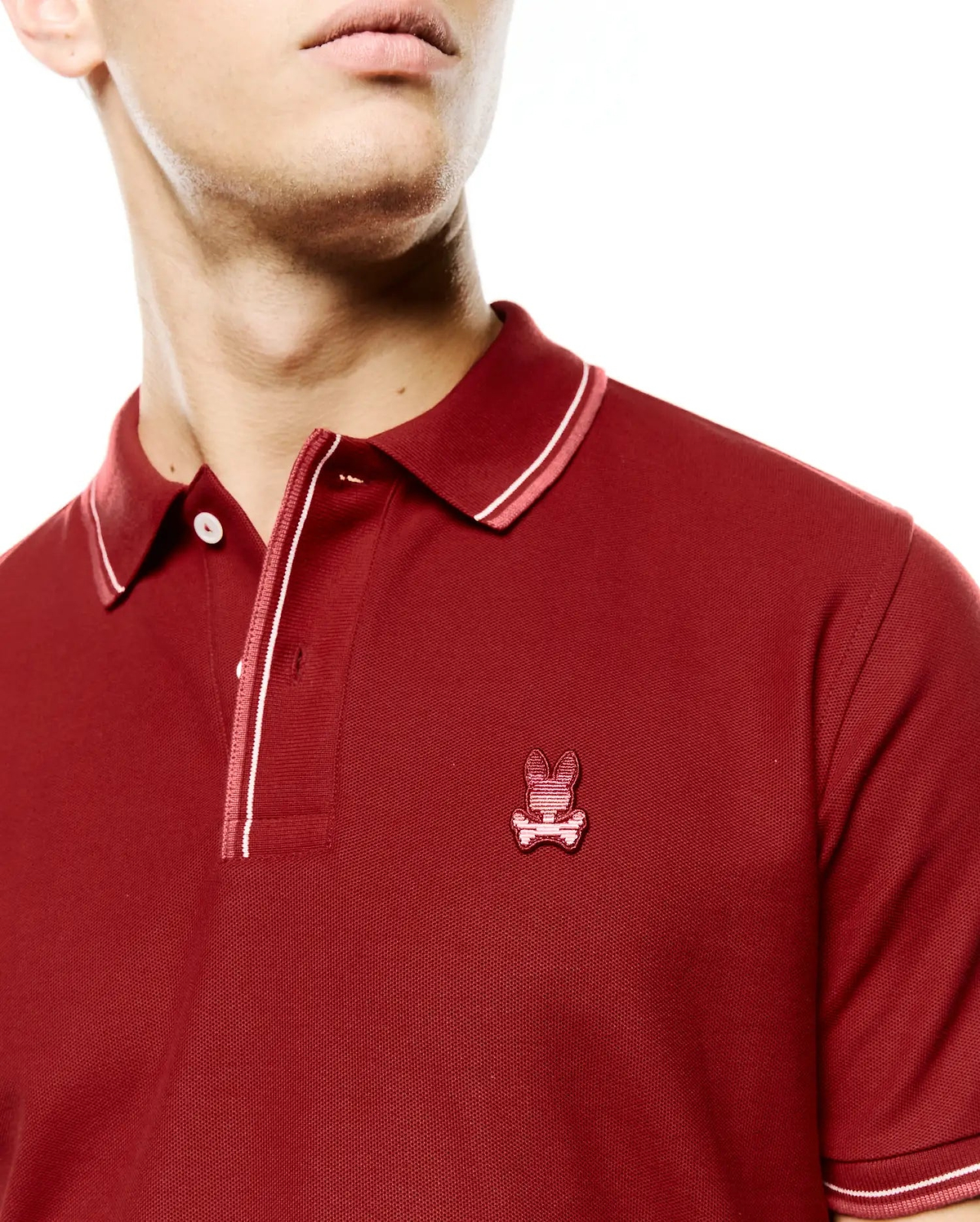 Men's Slade Pique Polo, a classic fit deep red pique polo featuring a small embroidered Psycho Bunny skull logo on the left chest and white with pink striped accents on the collar, placket, and cuffs. (2)