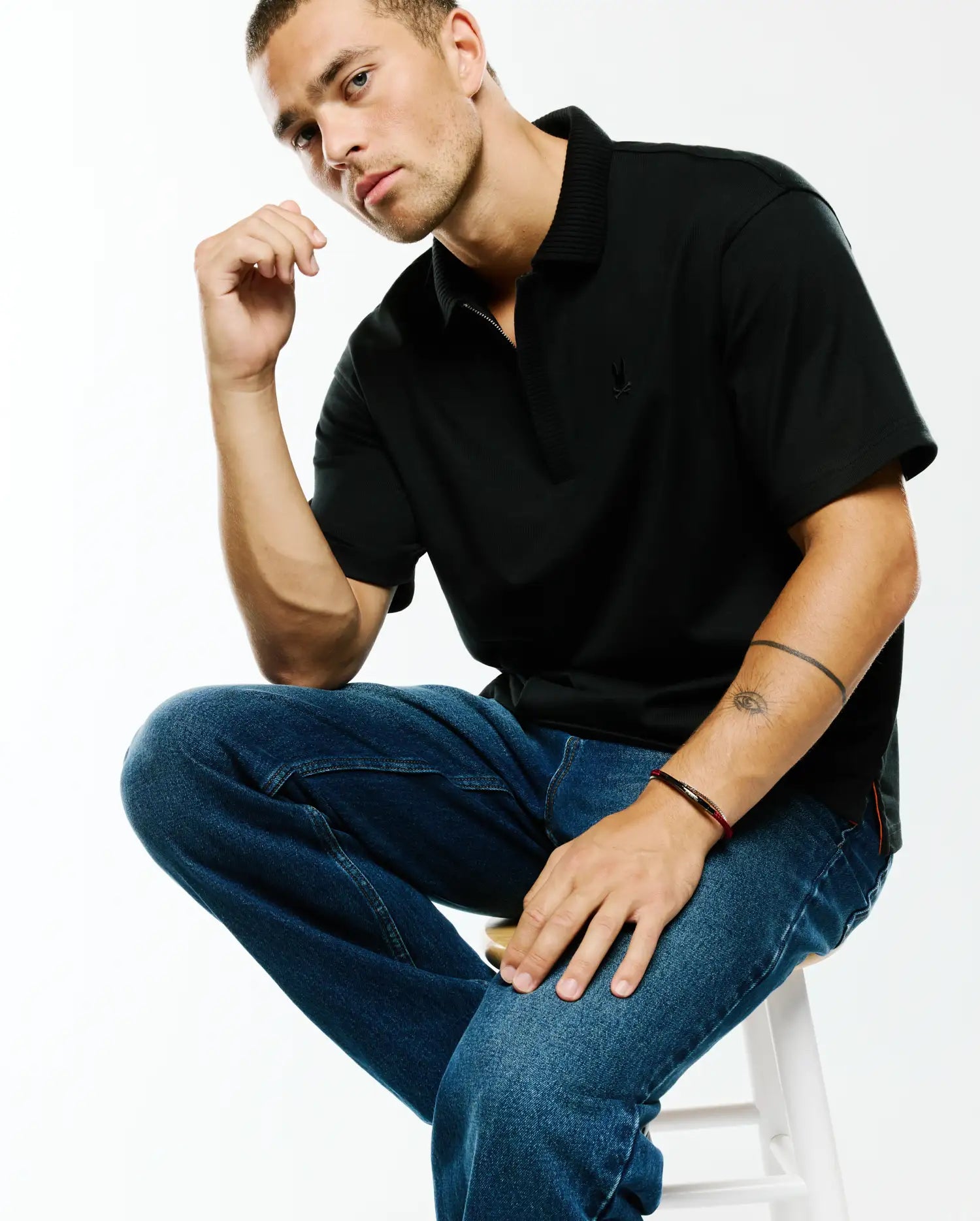 Men's Nolen Oversized Jersey Polo, a black oversized jersey polo with a ribbed collar, quarter-zip placket, small embroidered Psycho Bunny skull logo on the left chest, and subtle red side slit accents. (3)