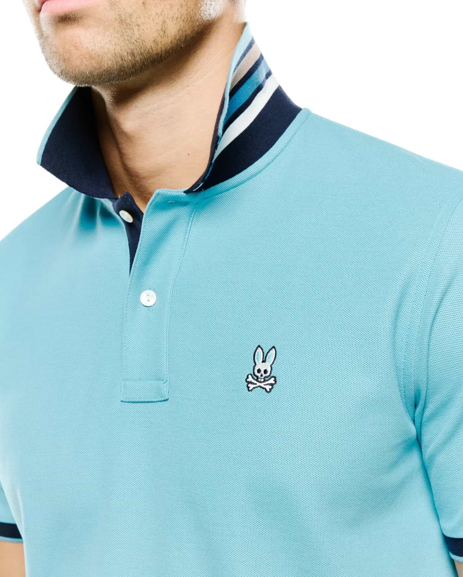 Men's Klaus Pique Polo, a light blue classic fit pique knit polo with a navy collar, two-button placket, and multi-striped inner collar, featuring the iconic embroidered Psycho Bunny skull logo on the left chest. (2)