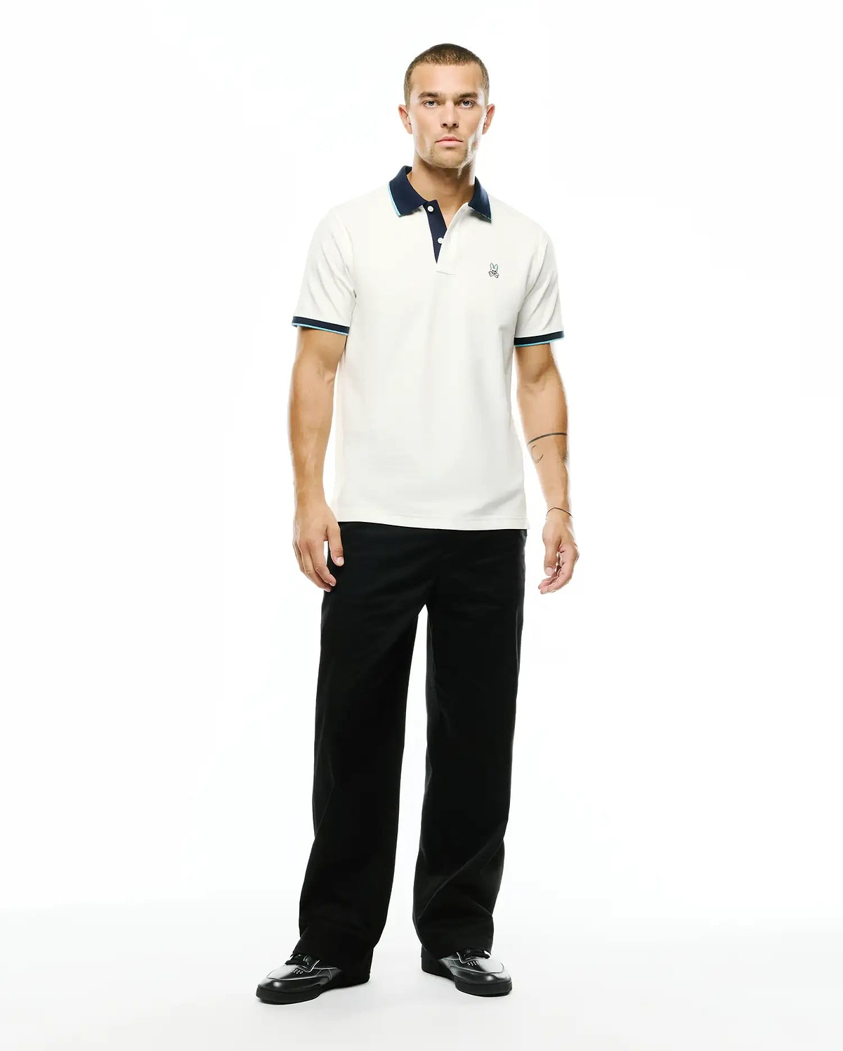 Men's Klaus Pique Polo in off-white, featuring a navy blue collar, light blue and navy striped ribbed cuffs, and a small embroidered Psycho Bunny skull logo on the left chest. (4)