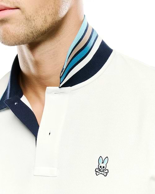 Men's Klaus Pique Polo in cream, featuring a classic fit, two-button placket, and a navy blue collar with multi-colored stripes in blues, white, and tan. The small, embroidered Psycho Bunny skull logo with blue ears is on the left chest. (2)