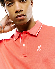 Mens Atley Pique Polo, vibrant coral, classic fit, pique knit. Features a two-button placket, ribbed collar with white tipping, and a small white embroidered Psycho Bunny skull logo on the left chest. (2)