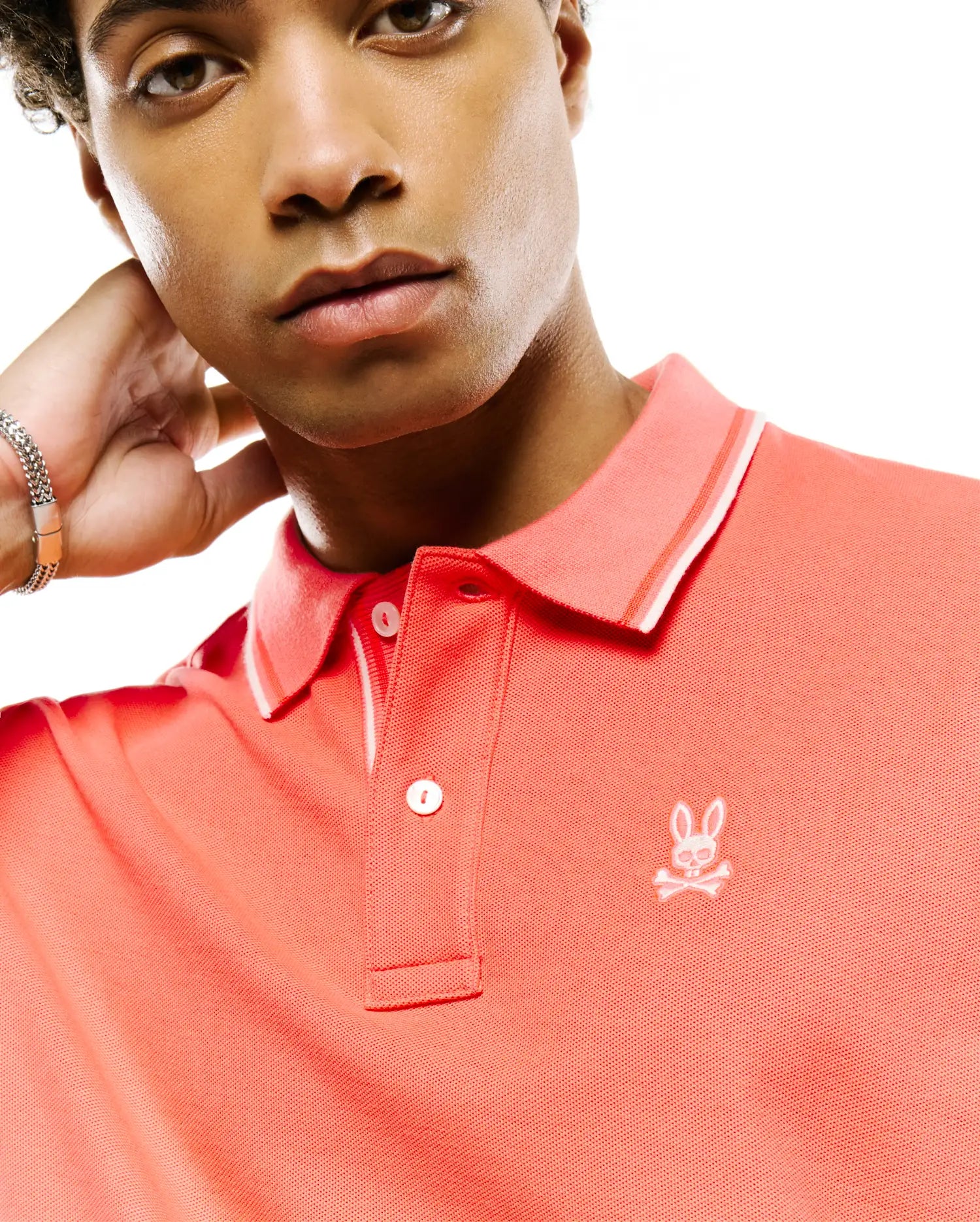 Mens Atley Pique Polo, vibrant coral, classic fit, pique knit. Features a two-button placket, ribbed collar with white tipping, and a small white embroidered Psycho Bunny skull logo on the left chest. (2)