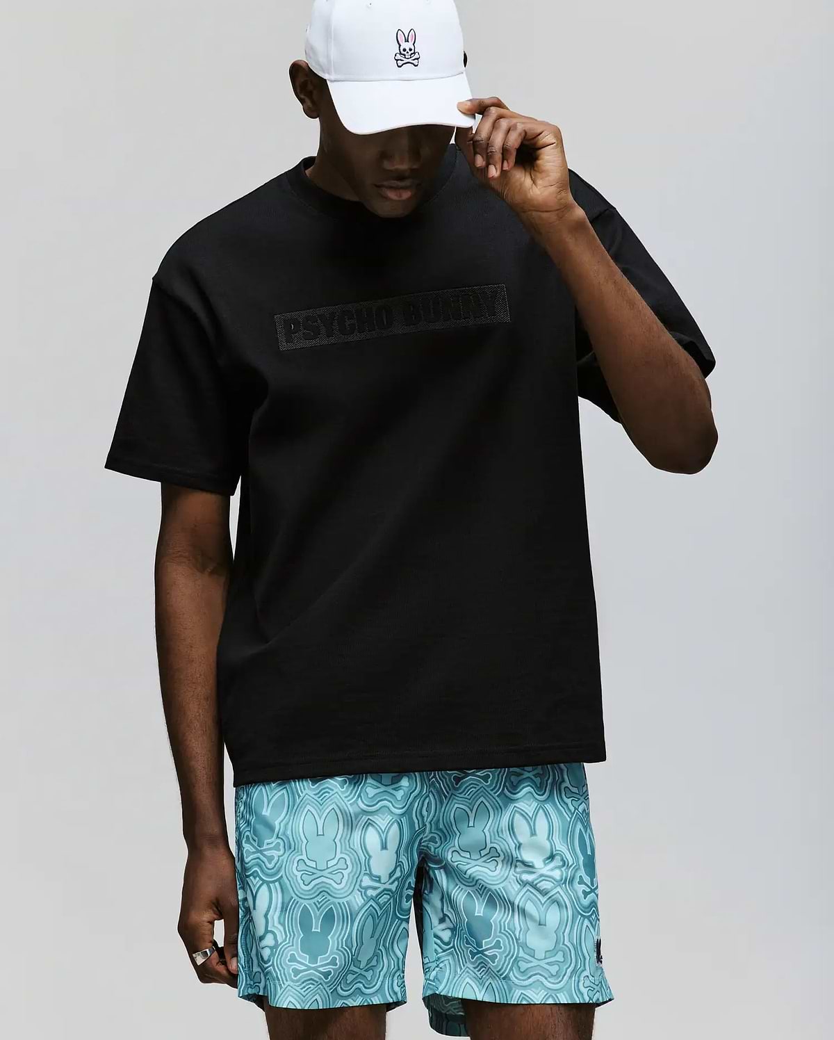 A person wearing the Psycho Bunny MENS ZAYN OVERSIZED GRAPHIC TEE (MT0200004), made from 100% cotton, paired with light blue bunny-patterned shorts and a white cap featuring a small bunny logo, stands against a plain background.