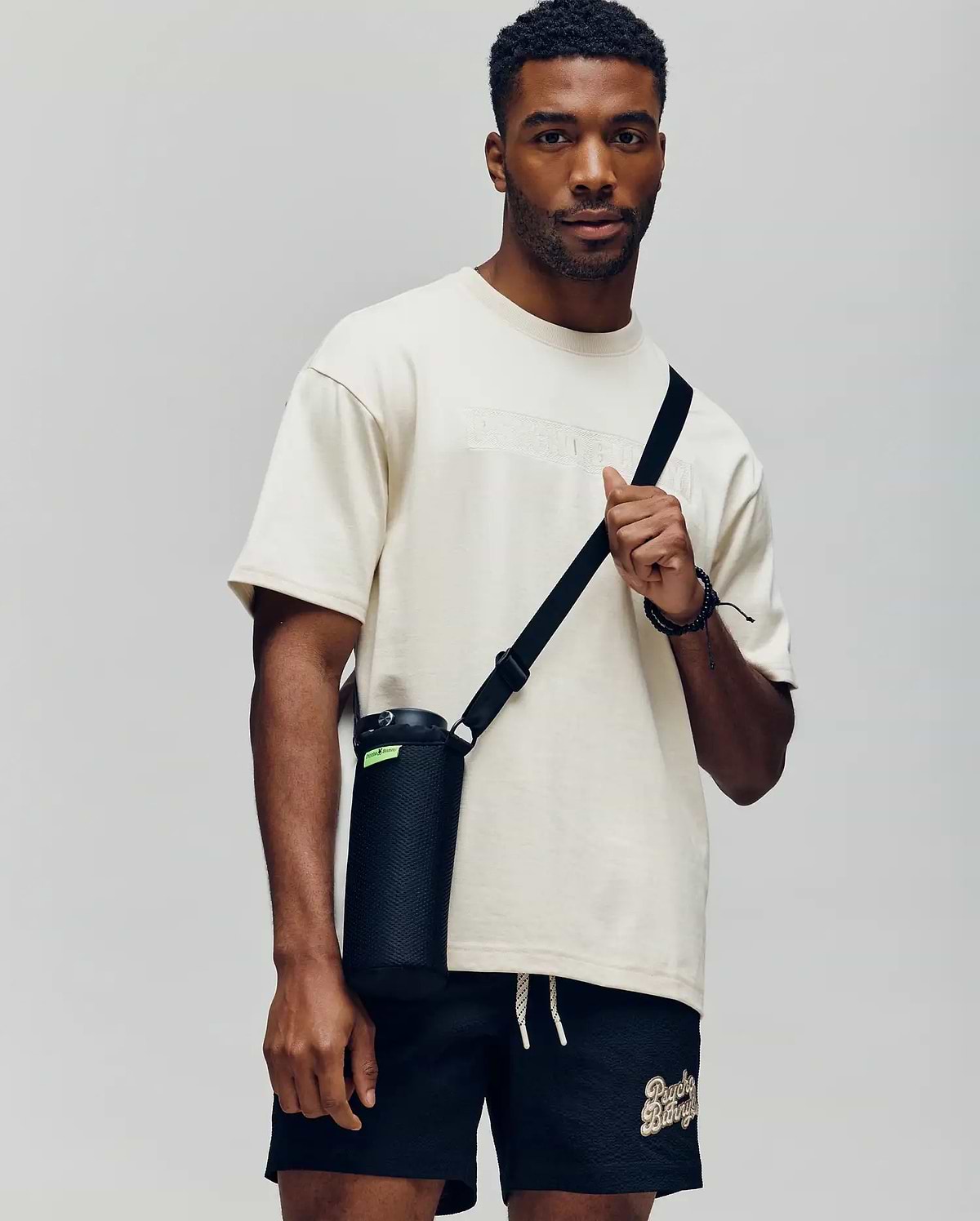 A man wears a Psycho Bunny MENS ZAYN OVERSIZED GRAPHIC TEE (MT0200004) in cream and black shorts, holding a black water bottle with a strap. The relaxed fit stands out against the plain, light background.