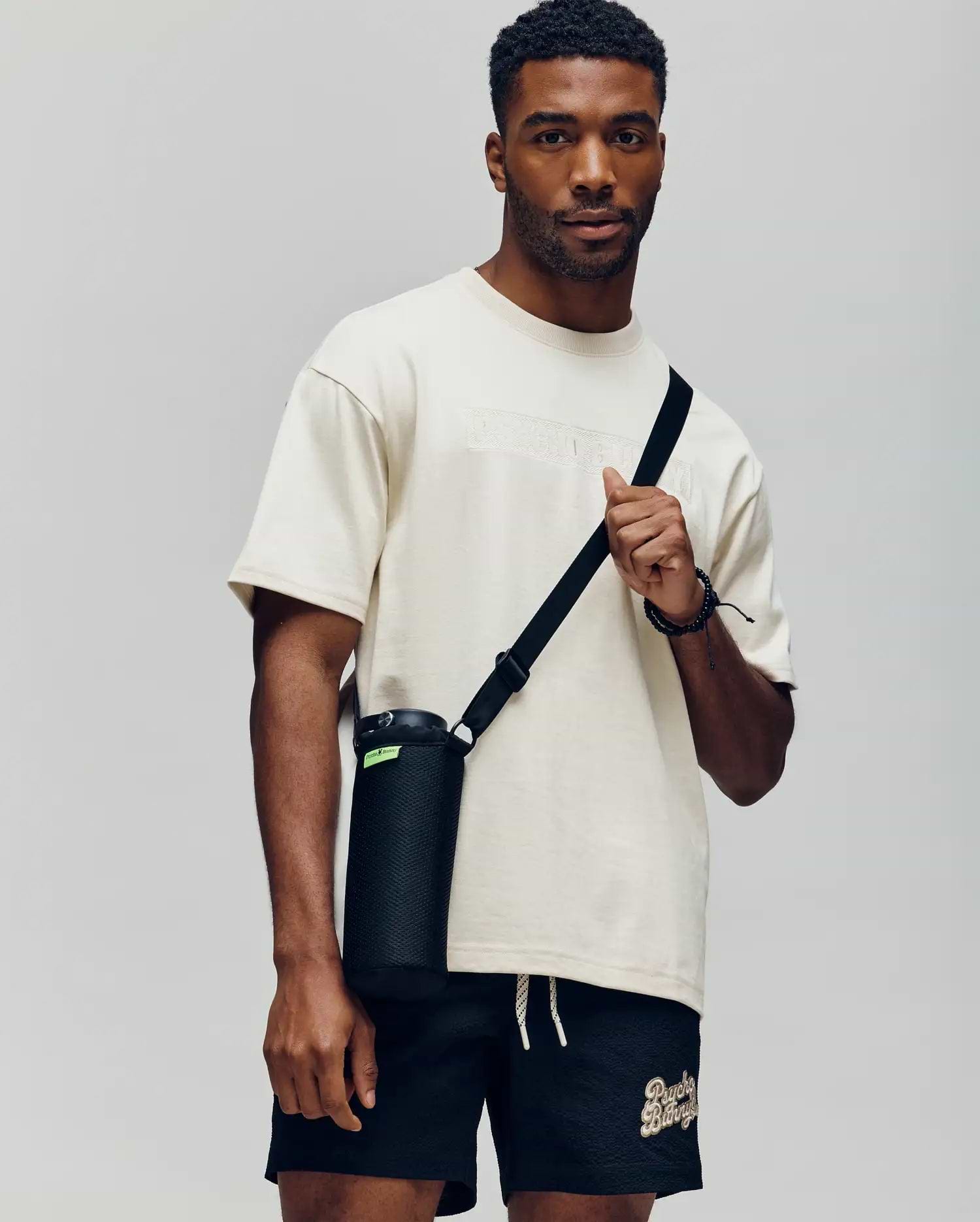 A man wears a Psycho Bunny MENS ZAYN OVERSIZED GRAPHIC TEE (MT0200004) in cream and black shorts, holding a black water bottle with a strap. The relaxed fit stands out against the plain, light background.