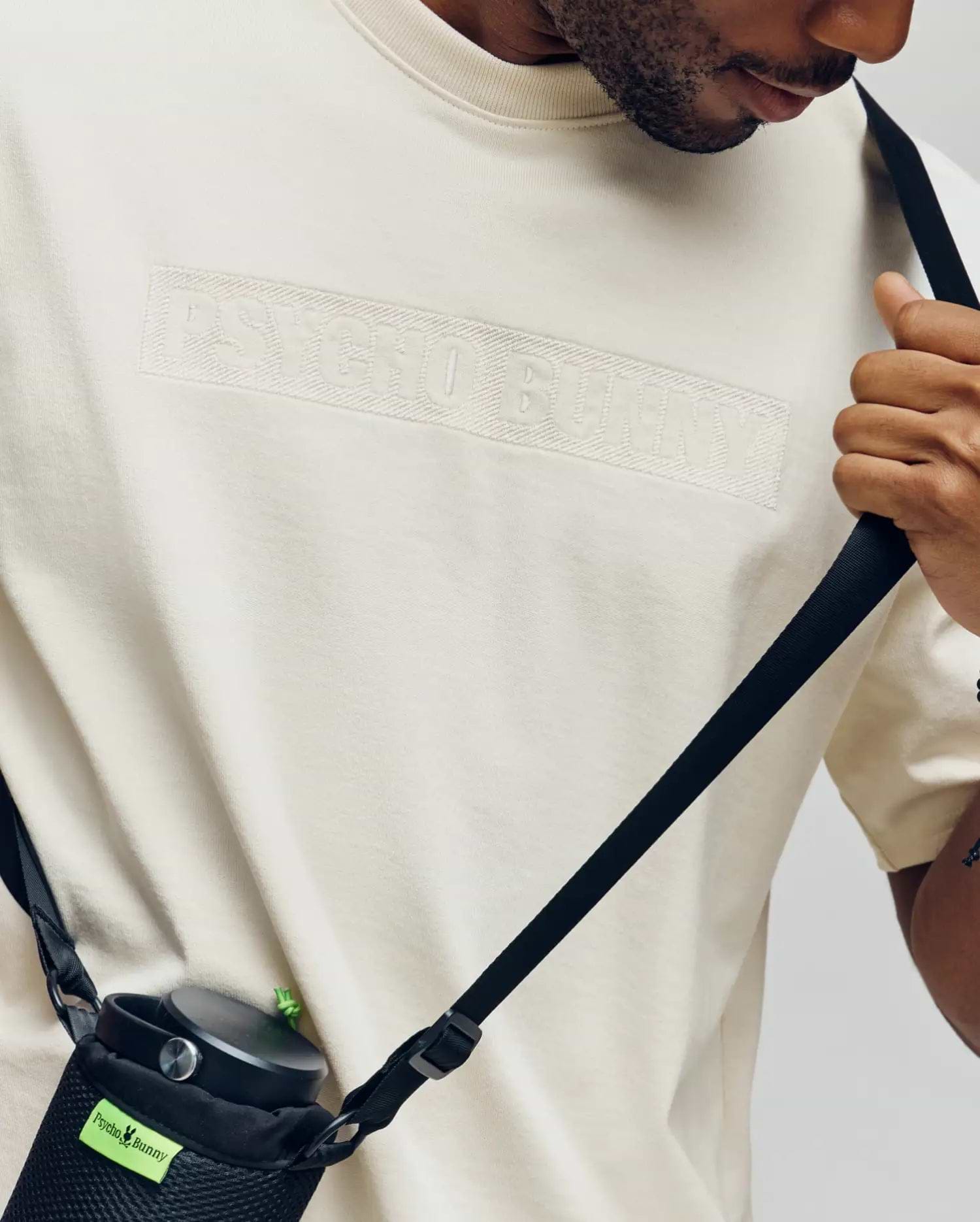 A person in a Psycho Bunny MENS ZAYN OVERSIZED GRAPHIC TEE (MT0200004) holds a black crossbody strap connected to a black water bottle holder with a green Psycho Bunny tag.