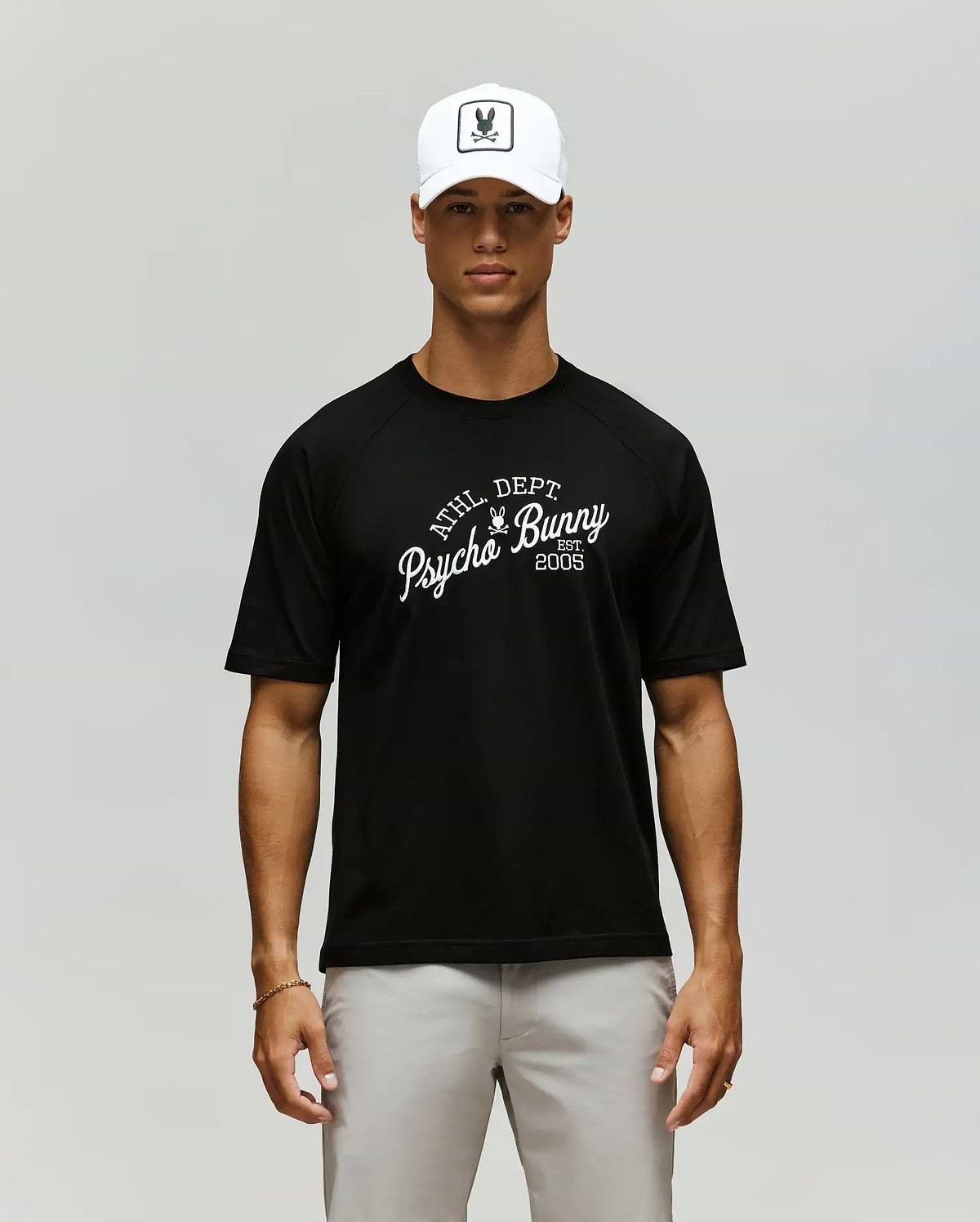 A man in a Psycho Bunny MENS MATTEO RAGLAN GRAPHIC TEE (MT0200008), white cap with bunny logo, and light gray pants stands against a plain, neutral background. (1)