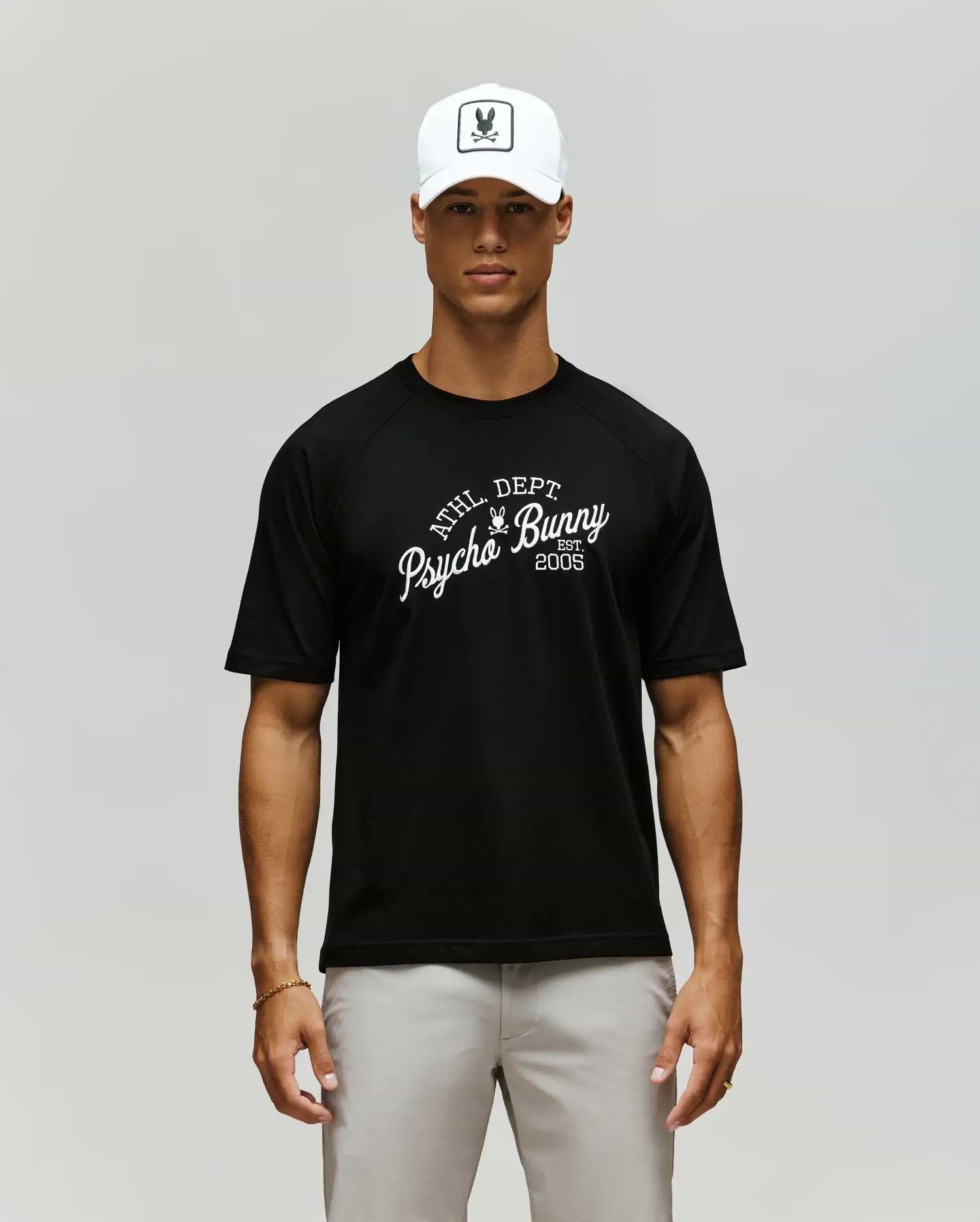 A man in a Psycho Bunny MENS MATTEO RAGLAN GRAPHIC TEE (MT0200008), white cap with bunny logo, and light gray pants stands against a plain, neutral background. (1)