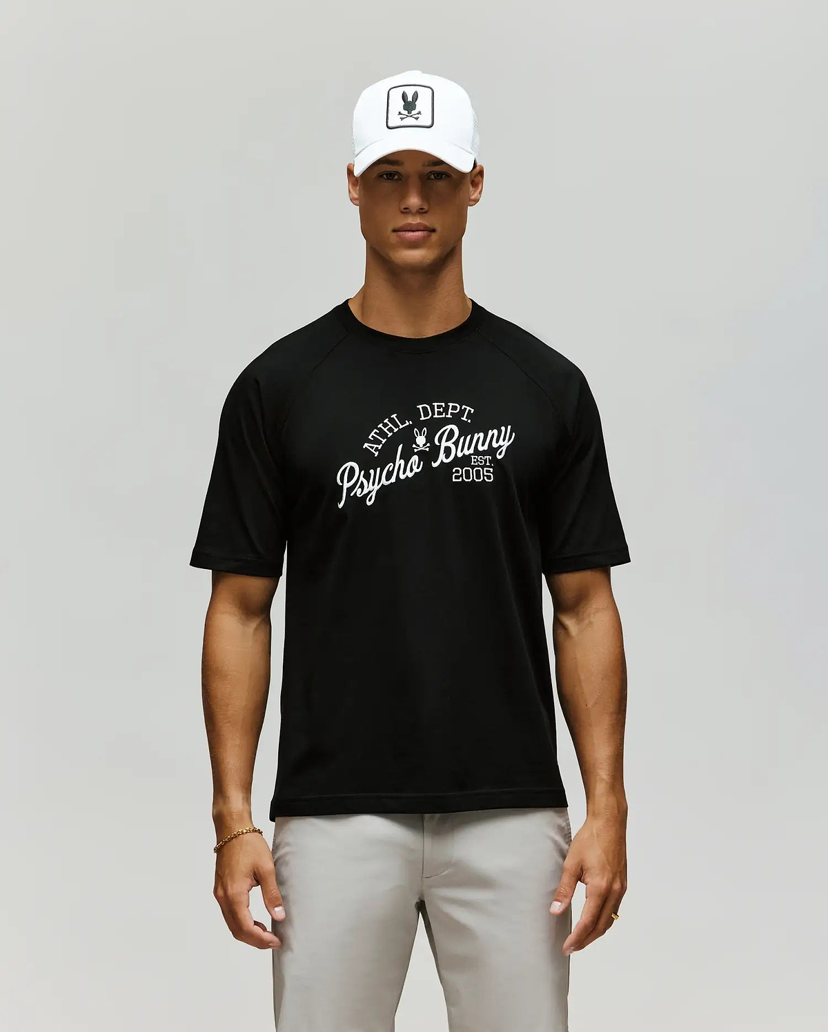 A man in a Psycho Bunny MENS MATTEO RAGLAN GRAPHIC TEE (MT0200008), white cap with bunny logo, and light gray pants stands against a plain, neutral background. (1)