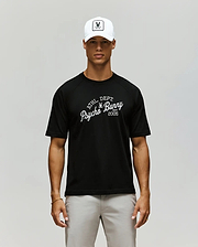 A man in a Psycho Bunny MENS MATTEO RAGLAN GRAPHIC TEE (MT0200008), white cap with bunny logo, and light gray pants stands against a plain, neutral background. (1)