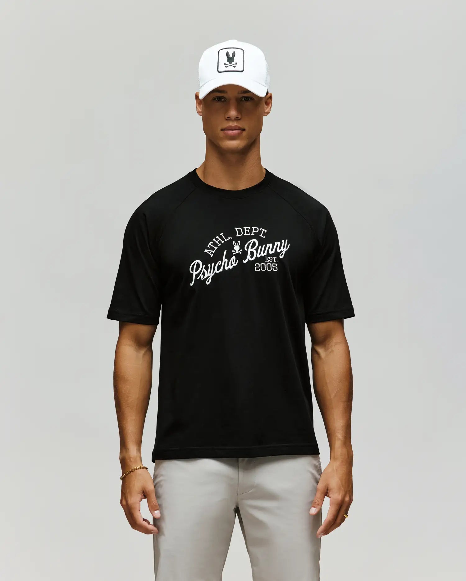 A man in a Psycho Bunny MENS MATTEO RAGLAN GRAPHIC TEE (MT0200008), white cap with bunny logo, and light gray pants stands against a plain, neutral background. (1)