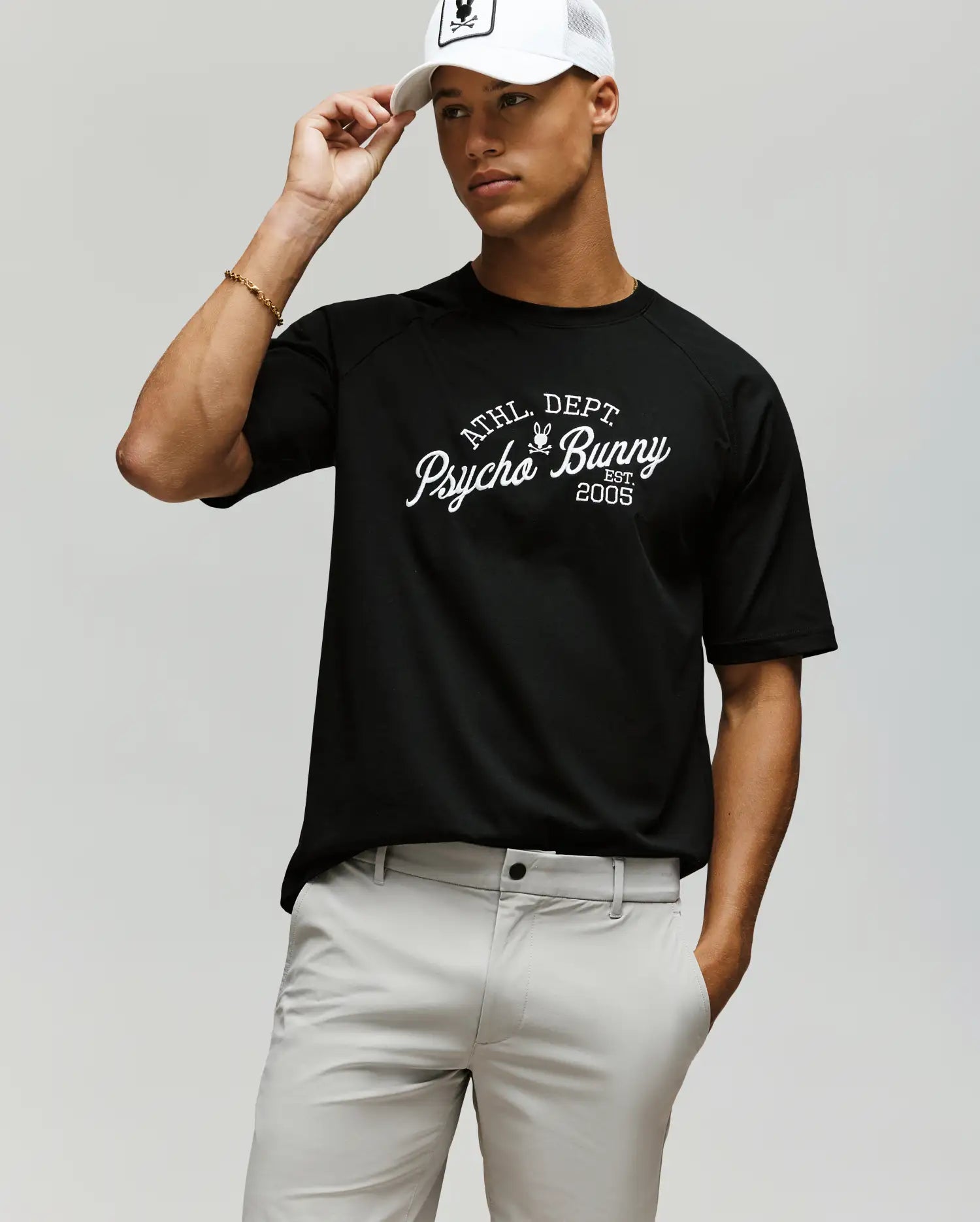 A person wearing a white cap, light gray pants, and a black Psycho Bunny MENS MATTEO RAGLAN GRAPHIC TEE (MT0200008) poses against a plain background, holding the brim of the cap with one hand. (2)