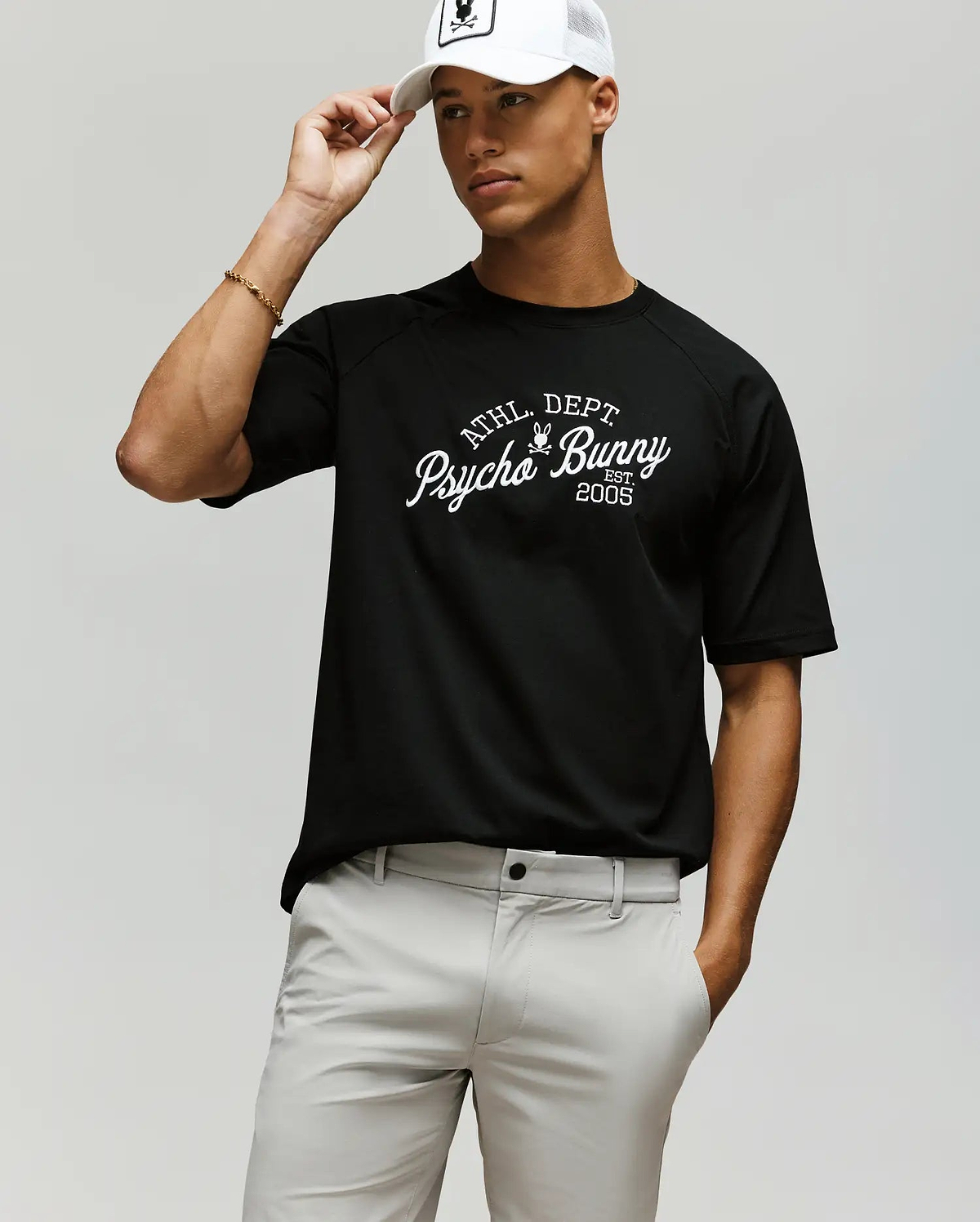 A person wearing a white cap, light gray pants, and a black Psycho Bunny MENS MATTEO RAGLAN GRAPHIC TEE (MT0200008) poses against a plain background, holding the brim of the cap with one hand. (2)