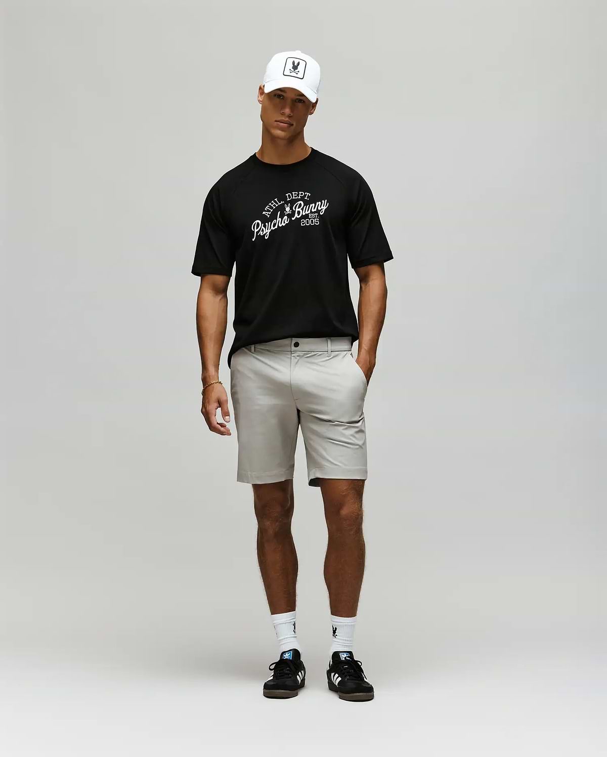 A man wearing a white baseball cap, light gray shorts, white socks, black sneakers, and a Psycho Bunny MENS MATTEO RAGLAN GRAPHIC TEE (MT0200008) stands against a plain light gray background. (4)