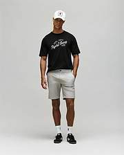 A man wearing a white baseball cap, light gray shorts, white socks, black sneakers, and a Psycho Bunny MENS MATTEO RAGLAN GRAPHIC TEE (MT0200008) stands against a plain light gray background. (4)