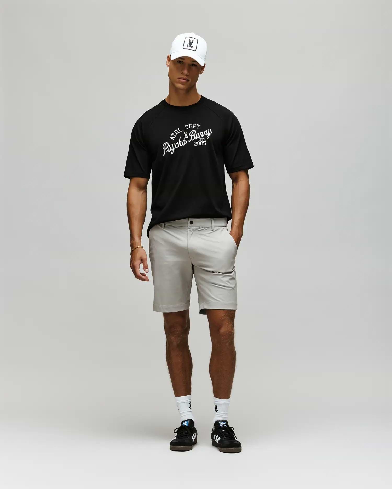 A man wearing a white baseball cap, light gray shorts, white socks, black sneakers, and a Psycho Bunny MENS MATTEO RAGLAN GRAPHIC TEE (MT0200008) stands against a plain light gray background. (4)