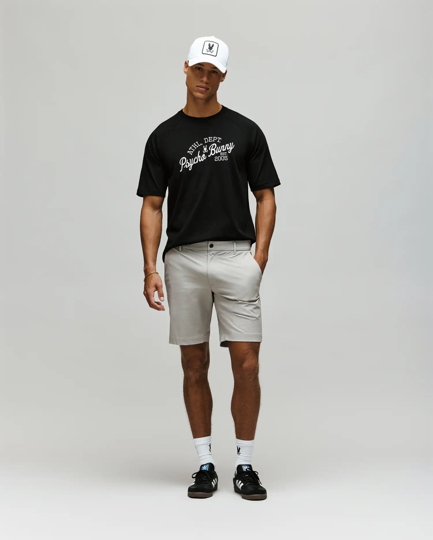 A man wearing a white baseball cap, light gray shorts, white socks, black sneakers, and a Psycho Bunny MENS MATTEO RAGLAN GRAPHIC TEE (MT0200008) stands against a plain light gray background. (4)