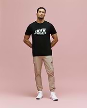 A man stands against a pink background wearing the Psycho Bunny MENS ARCHIVE GRADIANT BUNNY GRAPHIC TEE (MT0200029), paired with beige cargo pants and white high-top sneakers. He faces forward with his hands in his pockets. (4)