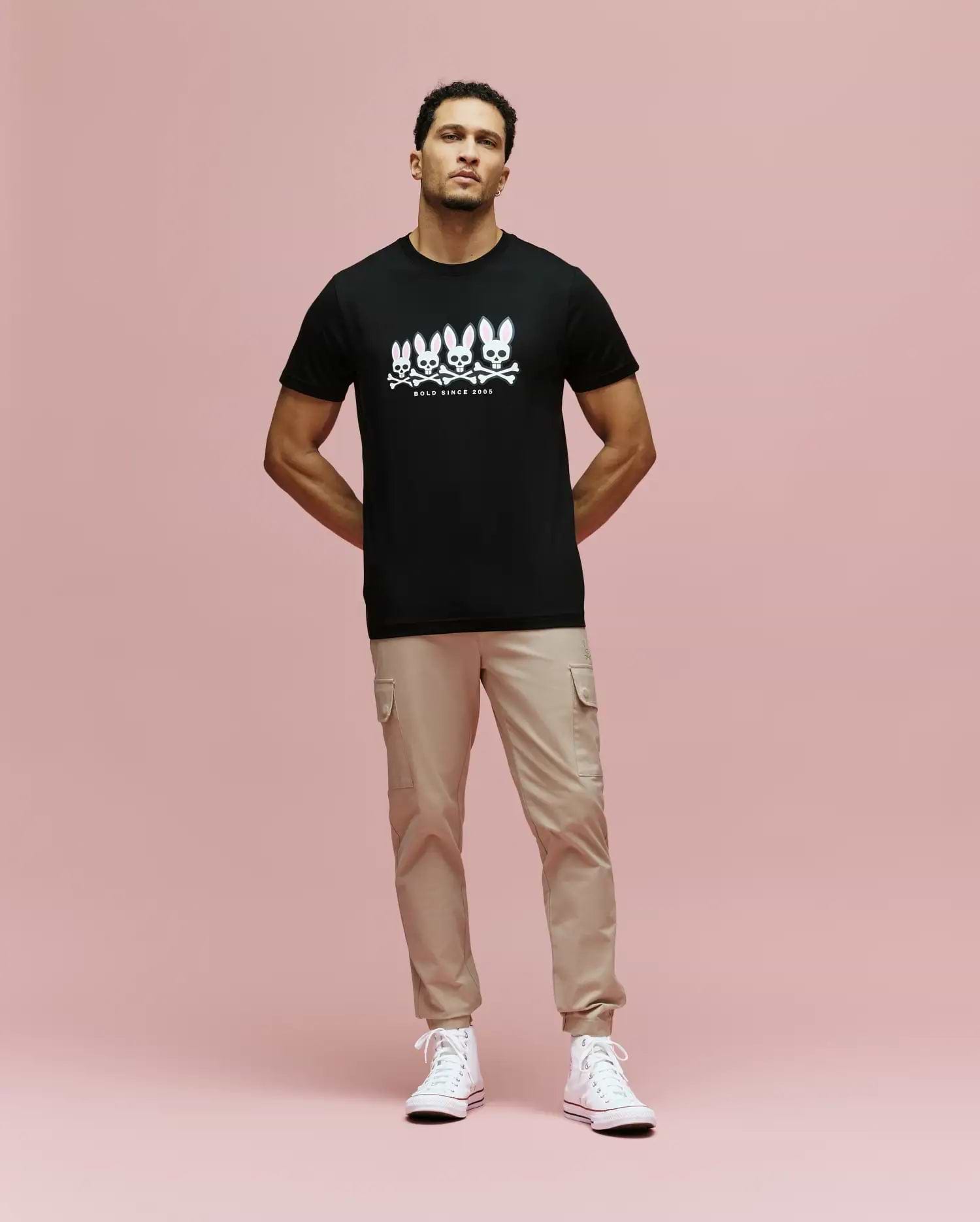 A man stands against a pink background wearing the Psycho Bunny MENS ARCHIVE GRADIANT BUNNY GRAPHIC TEE (MT0200029), paired with beige cargo pants and white high-top sneakers. He faces forward with his hands in his pockets. (4)