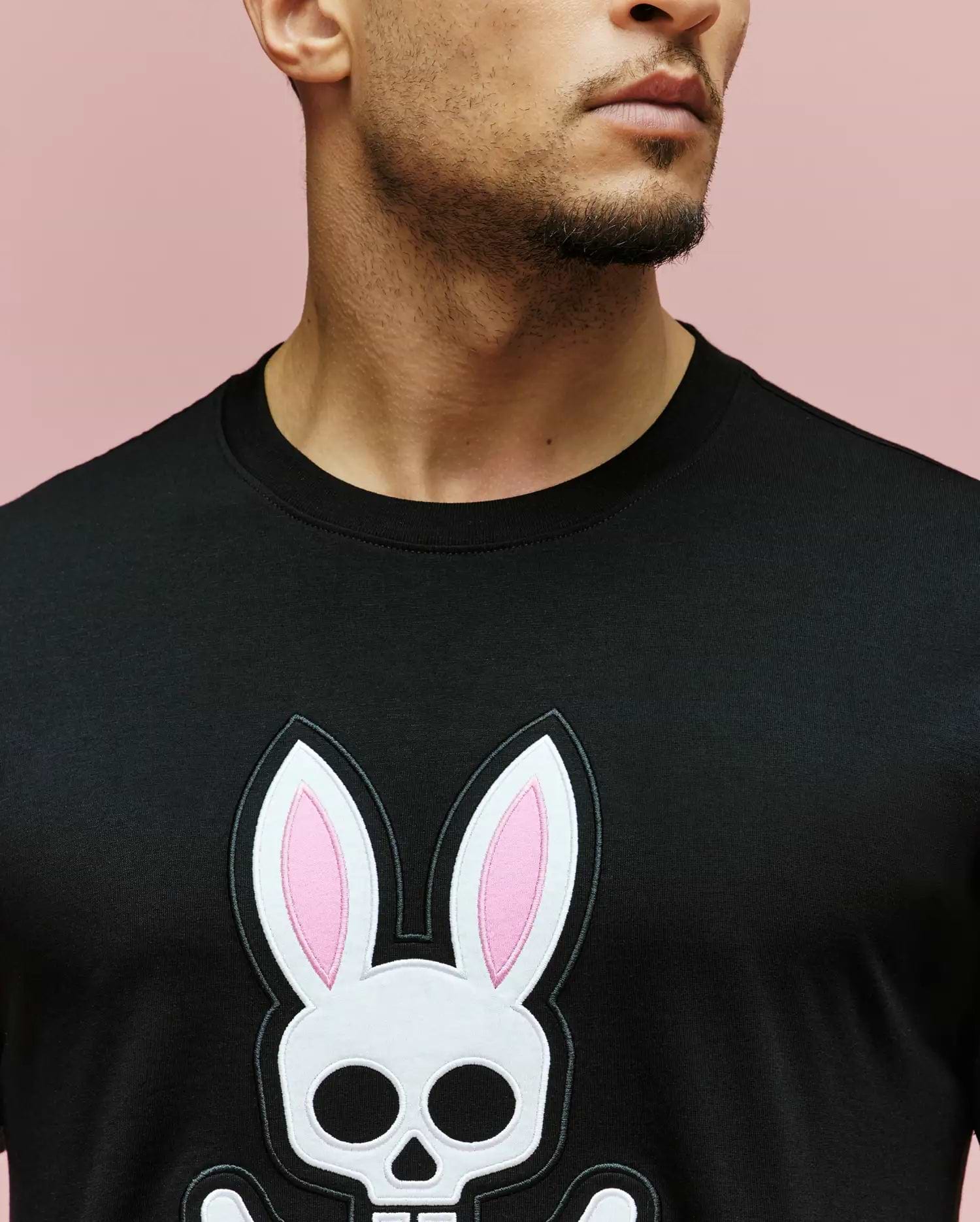 A man wears the Psycho Bunny MENS ARCHIVE BUNNY GRAPHIC TEE (MT0200031), a regular fit black shirt with a large graphic bunny—pink ears and skull face—on a pink background. Only his mouth, beard, and neck are visible in the image.
