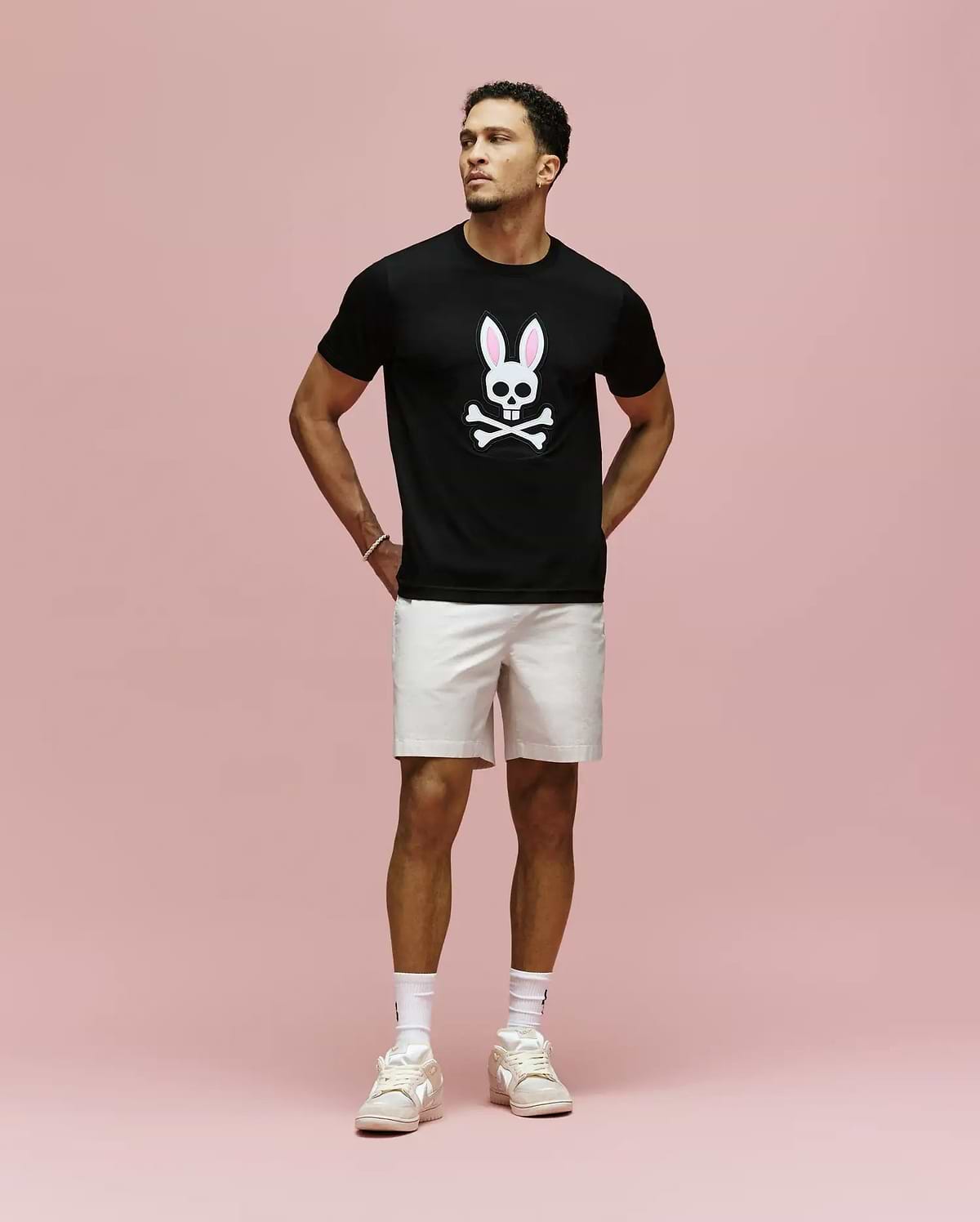 A man stands against a pink background wearing the Psycho Bunny MENS ARCHIVE BUNNY GRAPHIC TEE (MT0200031), black with an embroidered bunny and crossbones, paired with light shorts, white socks, and sneakers. Hands behind his back, he gazes to the side.