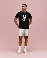 A man stands against a pink background wearing the Psycho Bunny MENS ARCHIVE BUNNY GRAPHIC TEE (MT0200031), black with an embroidered bunny and crossbones, paired with light shorts, white socks, and sneakers. Hands behind his back, he gazes to the side.