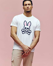 A man wears the Psycho Bunny MENS ARCHIVE BUNNY GRAPHIC TEE (MT0200031), a white rib knit crewneck with a skull and pink bunny ears graphic, standing against a plain pink background with his hands loosely clasped. (2)