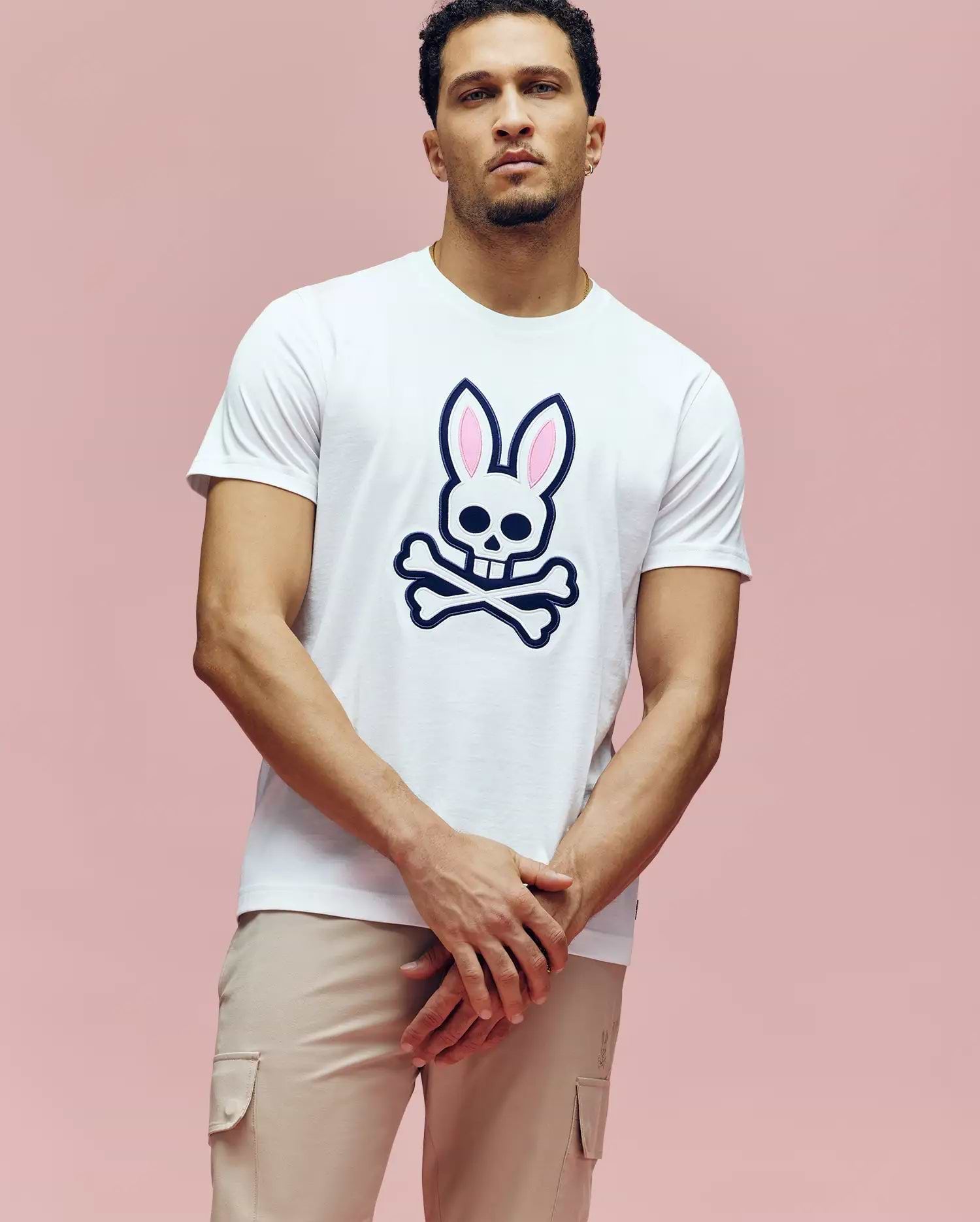 A man wears the Psycho Bunny MENS ARCHIVE BUNNY GRAPHIC TEE (MT0200031), a white rib knit crewneck with a skull and pink bunny ears graphic, standing against a plain pink background with his hands loosely clasped. (2)