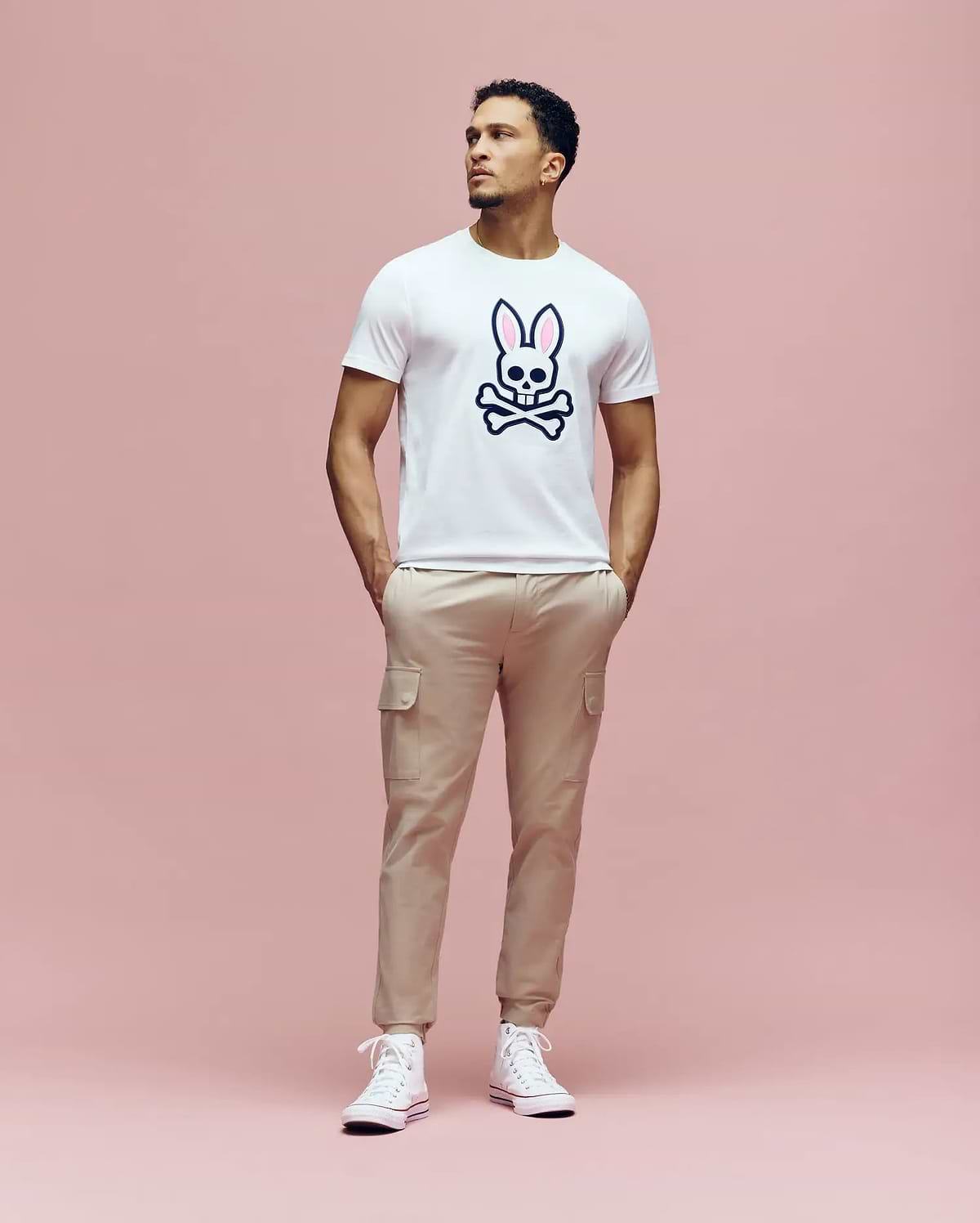 A man stands against a pink background, wearing the Psycho Bunny MENS ARCHIVE BUNNY GRAPHIC TEE (MT0200031) in white, with an embroidered bunny and crossbones graphic, paired with beige cargo pants and white sneakers. He looks up with his hands in his pockets. (4)