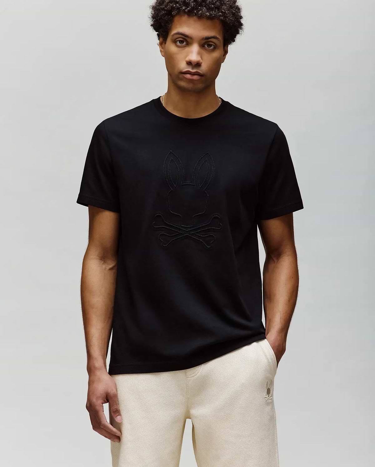 A man with short curly hair wears the Psycho Bunny MENS REMY GRAPHIC TEE (MT0200059) in black, made of soft Pima cotton with an embossed Bunny and crossbones logo, paired with light-colored pants against a light gray background.