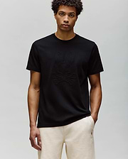 A man with short curly hair wears the Psycho Bunny MENS REMY GRAPHIC TEE (MT0200059) in black, made of soft Pima cotton with an embossed Bunny and crossbones logo, paired with light-colored pants against a light gray background.