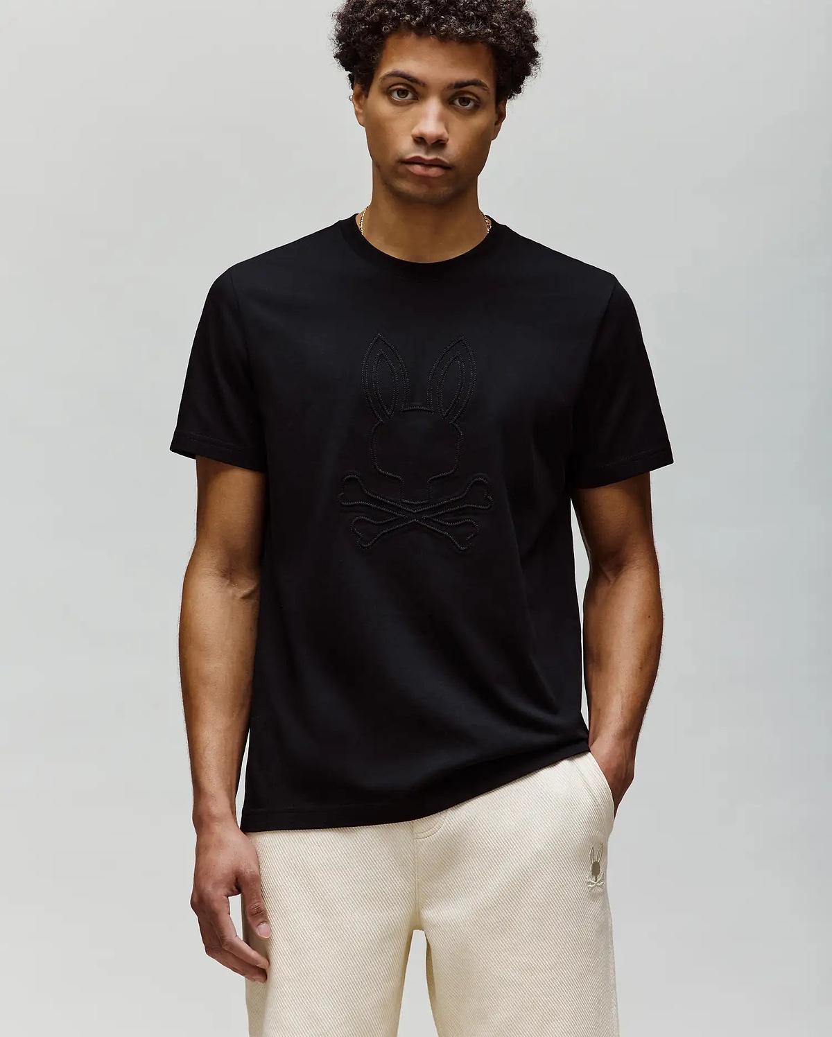 A man with short curly hair wears the Psycho Bunny MENS REMY GRAPHIC TEE (MT0200059) in black, made of soft Pima cotton with an embossed Bunny and crossbones logo, paired with light-colored pants against a light gray background.