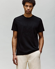 A man with short curly hair wears the Psycho Bunny MENS REMY GRAPHIC TEE (MT0200059) in black, made of soft Pima cotton with an embossed Bunny and crossbones logo, paired with light-colored pants against a light gray background.