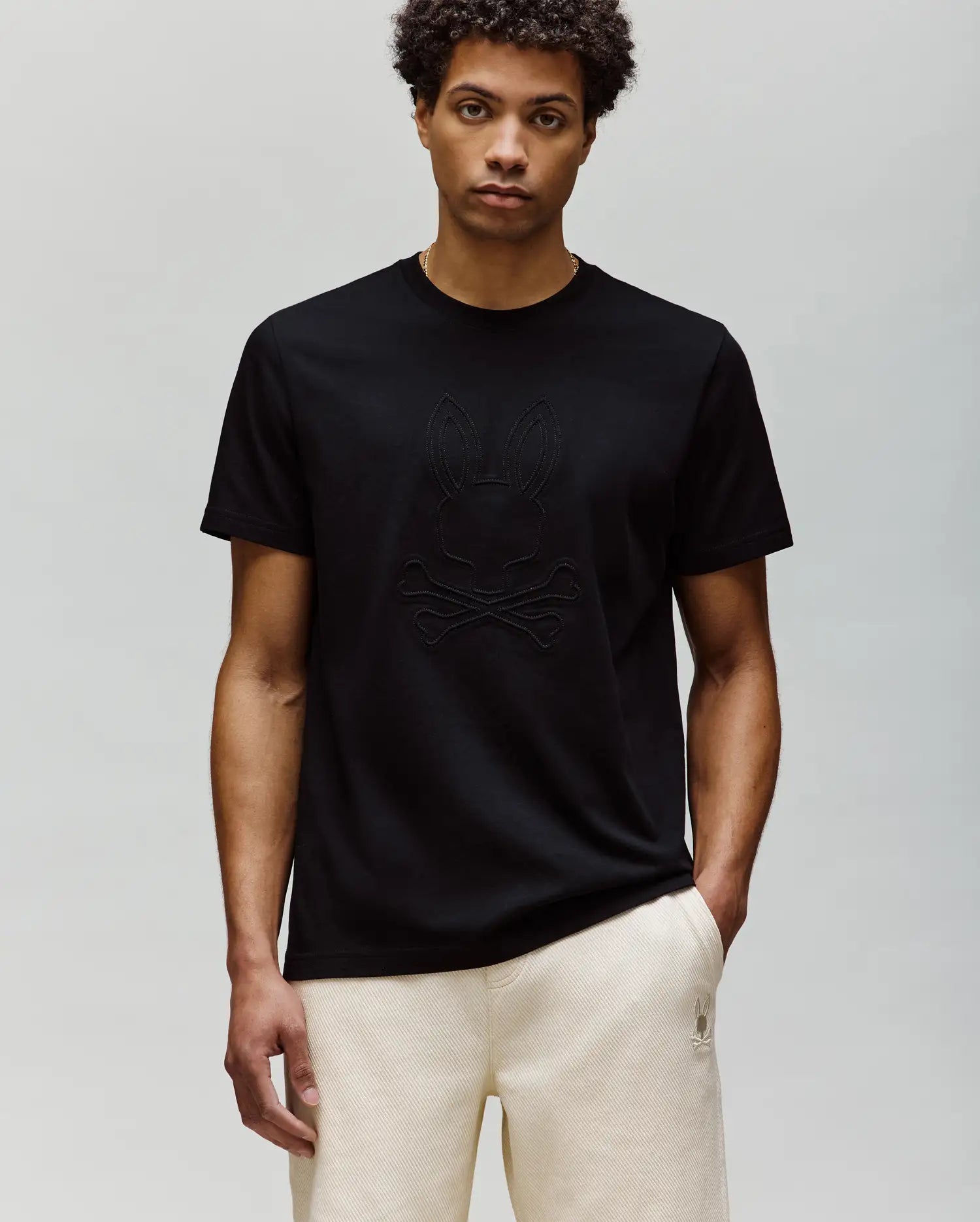 A man with short curly hair wears the Psycho Bunny MENS REMY GRAPHIC TEE (MT0200059) in black, made of soft Pima cotton with an embossed Bunny and crossbones logo, paired with light-colored pants against a light gray background.