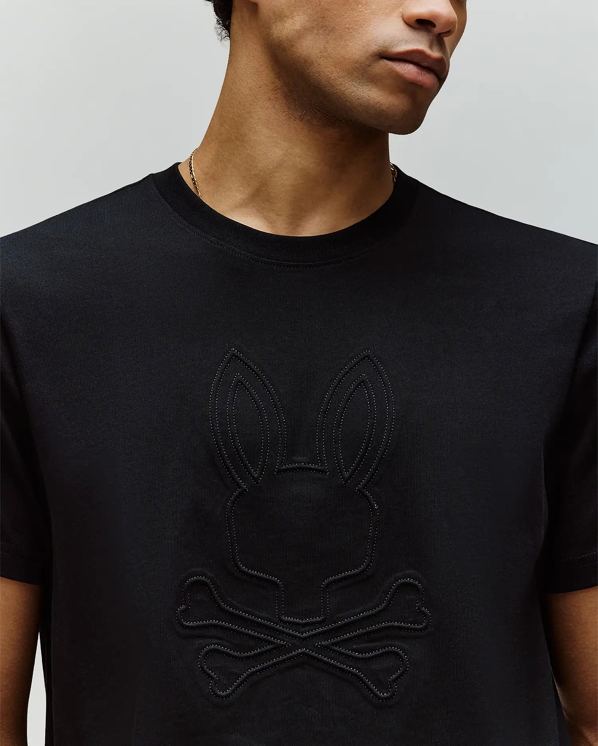 A man wears the Psycho Bunny MENS REMY GRAPHIC TEE (MT0200059), a black Pima cotton shirt featuring an embossed bunny logo above crossed bones, shown from the shoulders up.