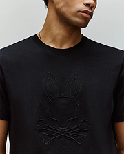 A man wears the Psycho Bunny MENS REMY GRAPHIC TEE (MT0200059), a black Pima cotton shirt featuring an embossed bunny logo above crossed bones, shown from the shoulders up.