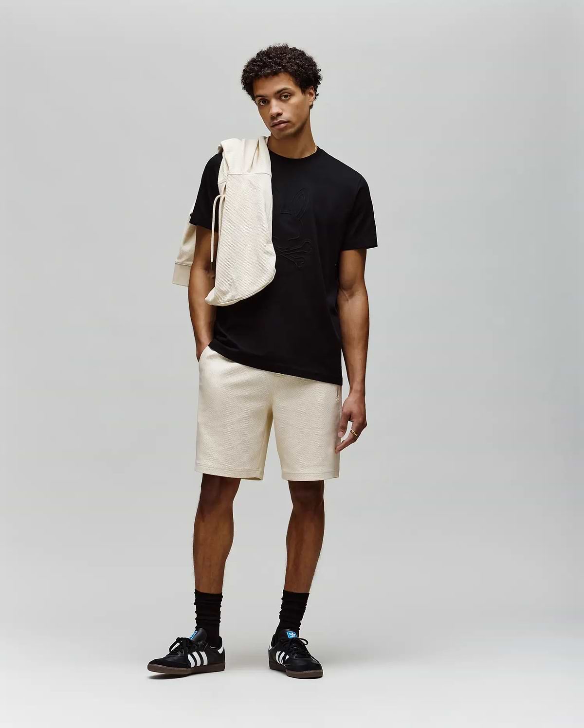 A young man stands against a plain background wearing a black MENS REMY GRAPHIC TEE - MT0200059 by Psycho Bunny, beige shorts, black socks, black Adidas sneakers, and a beige sweatshirt draped over his shoulder.