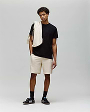 A young man stands against a plain background wearing a black MENS REMY GRAPHIC TEE - MT0200059 by Psycho Bunny, beige shorts, black socks, black Adidas sneakers, and a beige sweatshirt draped over his shoulder.
