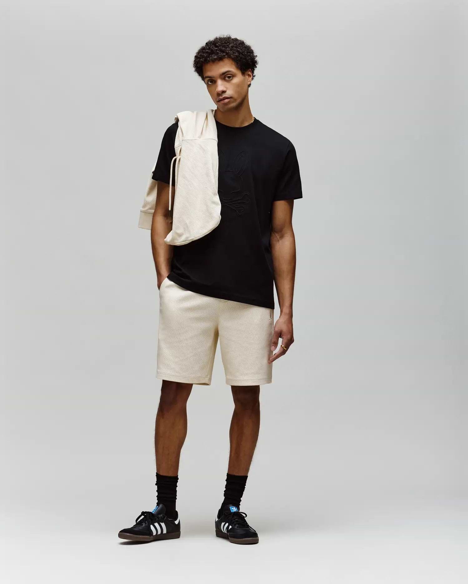 A young man stands against a plain background wearing a black MENS REMY GRAPHIC TEE - MT0200059 by Psycho Bunny, beige shorts, black socks, black Adidas sneakers, and a beige sweatshirt draped over his shoulder.
