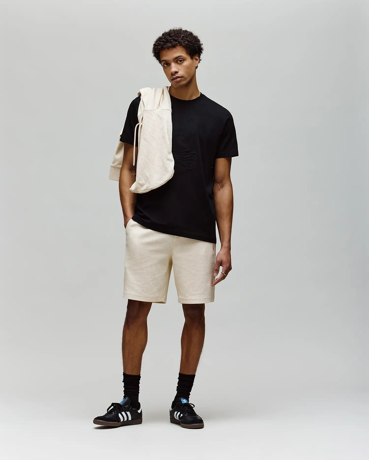 A young man stands against a plain background wearing a black MENS REMY GRAPHIC TEE - MT0200059 by Psycho Bunny, beige shorts, black socks, black Adidas sneakers, and a beige sweatshirt draped over his shoulder.