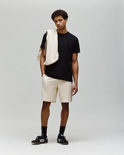 A young man stands against a plain background wearing a black MENS REMY GRAPHIC TEE - MT0200059 by Psycho Bunny, beige shorts, black socks, black Adidas sneakers, and a beige sweatshirt draped over his shoulder.