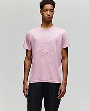 A young man wears the Psycho Bunny MENS REMY GRAPHIC TEE - MT0200059 in light pink, featuring the signature Bunny and crossbones logo on the chest, paired with black pants and a silver bracelet against a plain light background. (2)