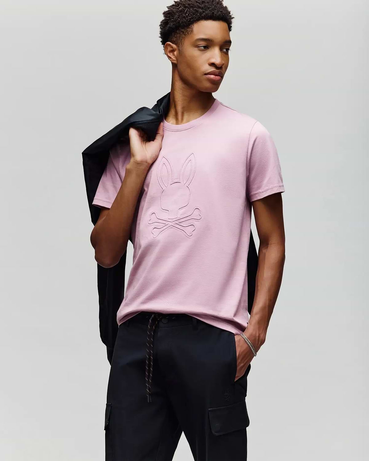 A young man poses against a light gray background, holding a black jacket over his shoulder and wearing Psycho Bunny's MENS REMY GRAPHIC TEE (MT0200059) in light pink, featuring an embossed Bunny logo and crossbones design, paired with black pants. (4)