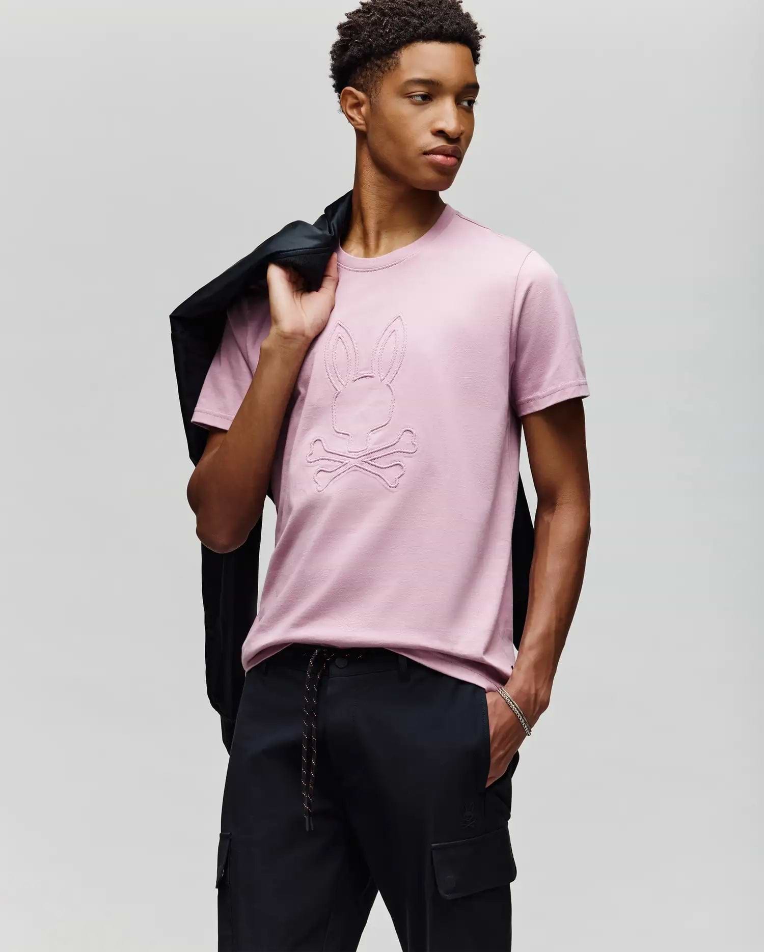 A young man poses against a light gray background, holding a black jacket over his shoulder and wearing Psycho Bunny's MENS REMY GRAPHIC TEE (MT0200059) in light pink, featuring an embossed Bunny logo and crossbones design, paired with black pants. (4)