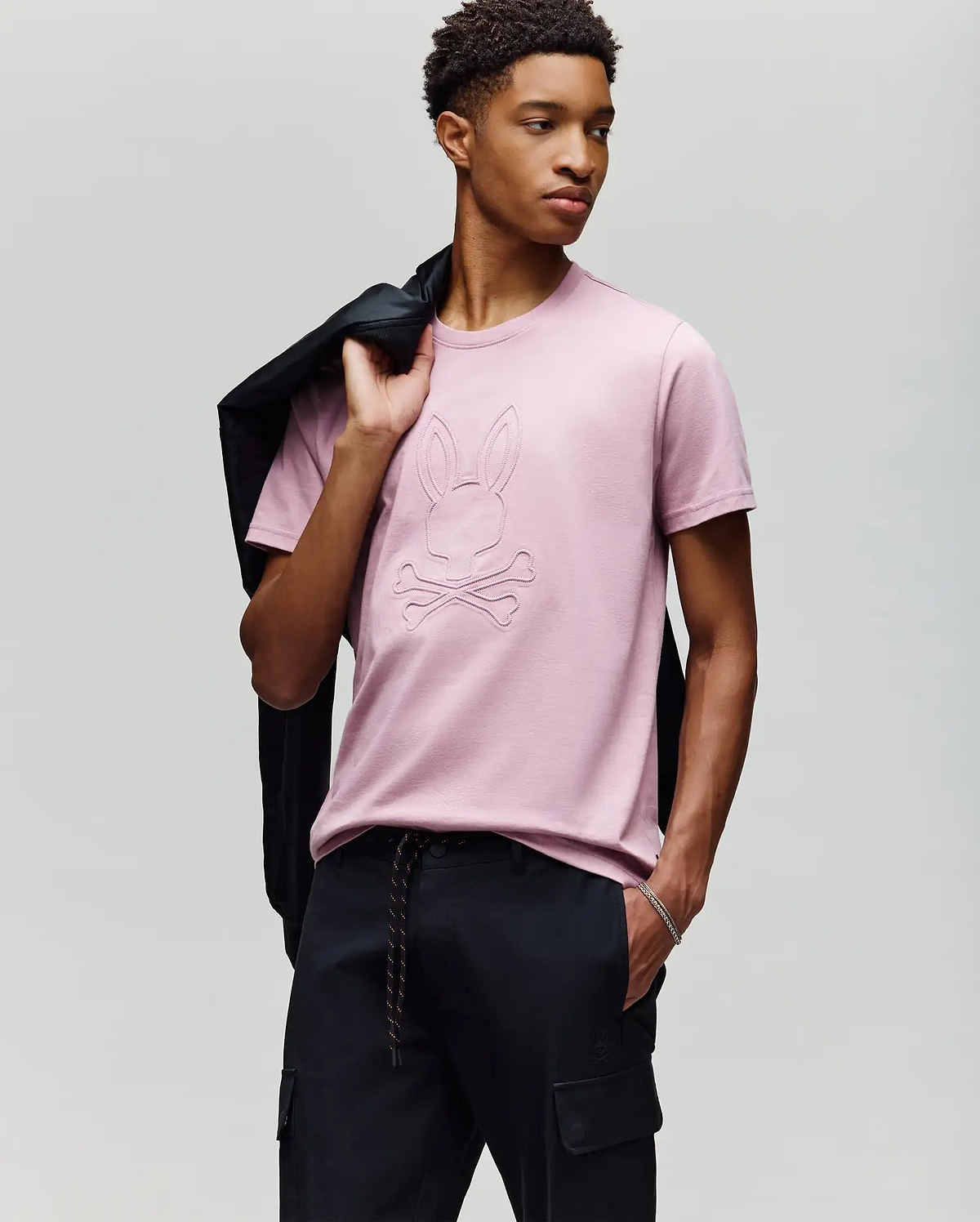 A young man poses against a light gray background, holding a black jacket over his shoulder and wearing Psycho Bunny's MENS REMY GRAPHIC TEE (MT0200059) in light pink, featuring an embossed Bunny logo and crossbones design, paired with black pants. (4)
