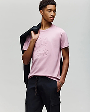 A young man poses against a light gray background, holding a black jacket over his shoulder and wearing Psycho Bunny's MENS REMY GRAPHIC TEE (MT0200059) in light pink, featuring an embossed Bunny logo and crossbones design, paired with black pants. (4)